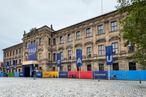Schlossplatz zeigt Schloss mit Flaggen und Bauzaunbespannung mit dem neuen FAU-Logo