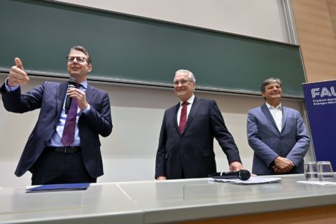 Three men stand in front of a blackboard in a lecture hall.