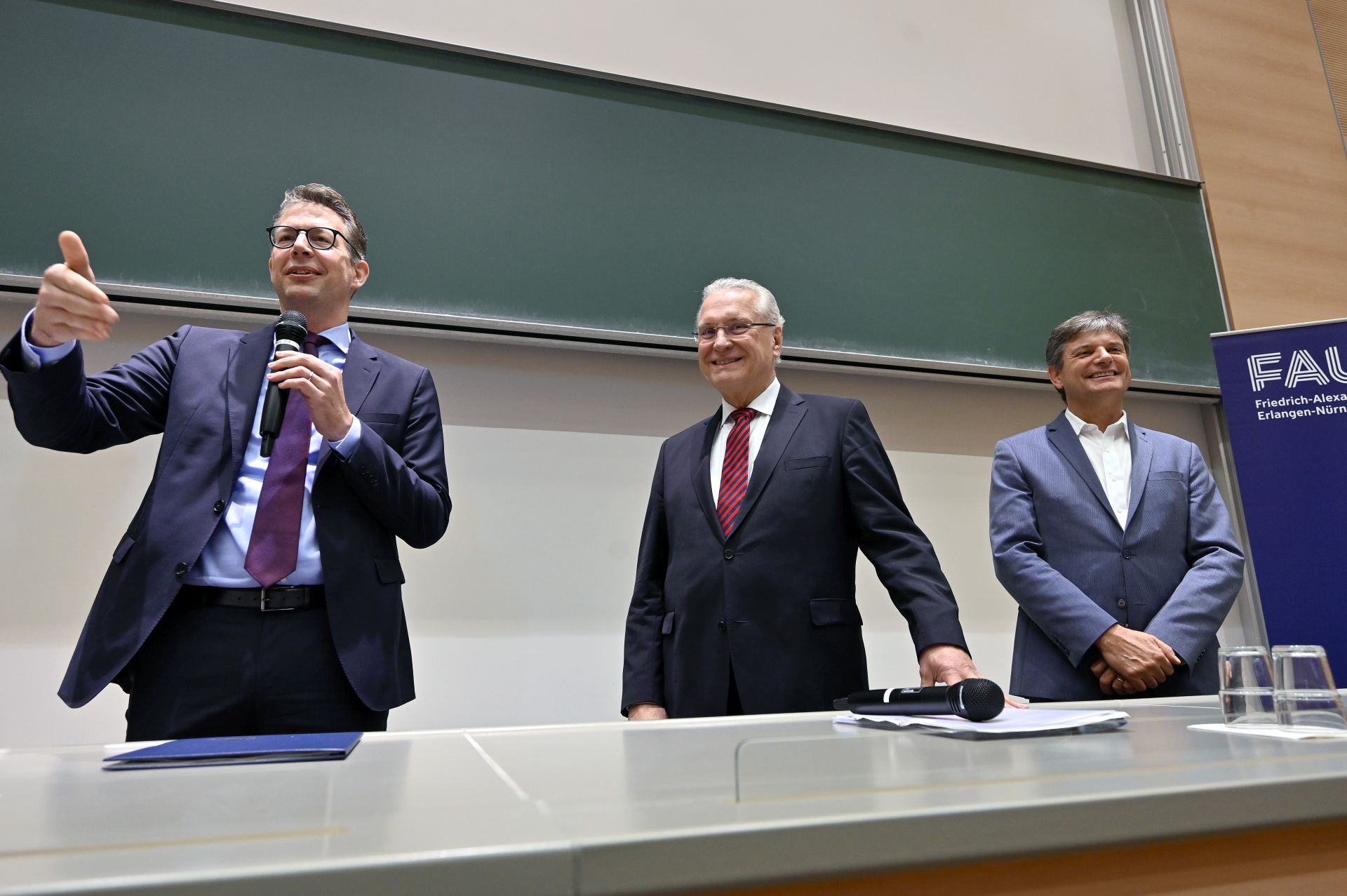 Three men stand in front of a blackboard in a lecture hall.