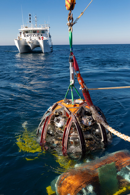 A DU, rolled up over a LOM which is installed on the DU base anchor, is prepared for deployment on the seafloor. In the background is the vessel from which the ROV is operated.