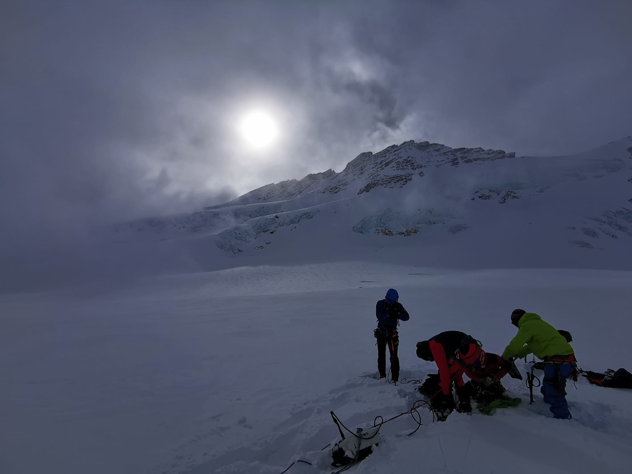 Researchers in protective winter clothing working on an ice field. The sun is behind the clouds, but you can still see it shining.
