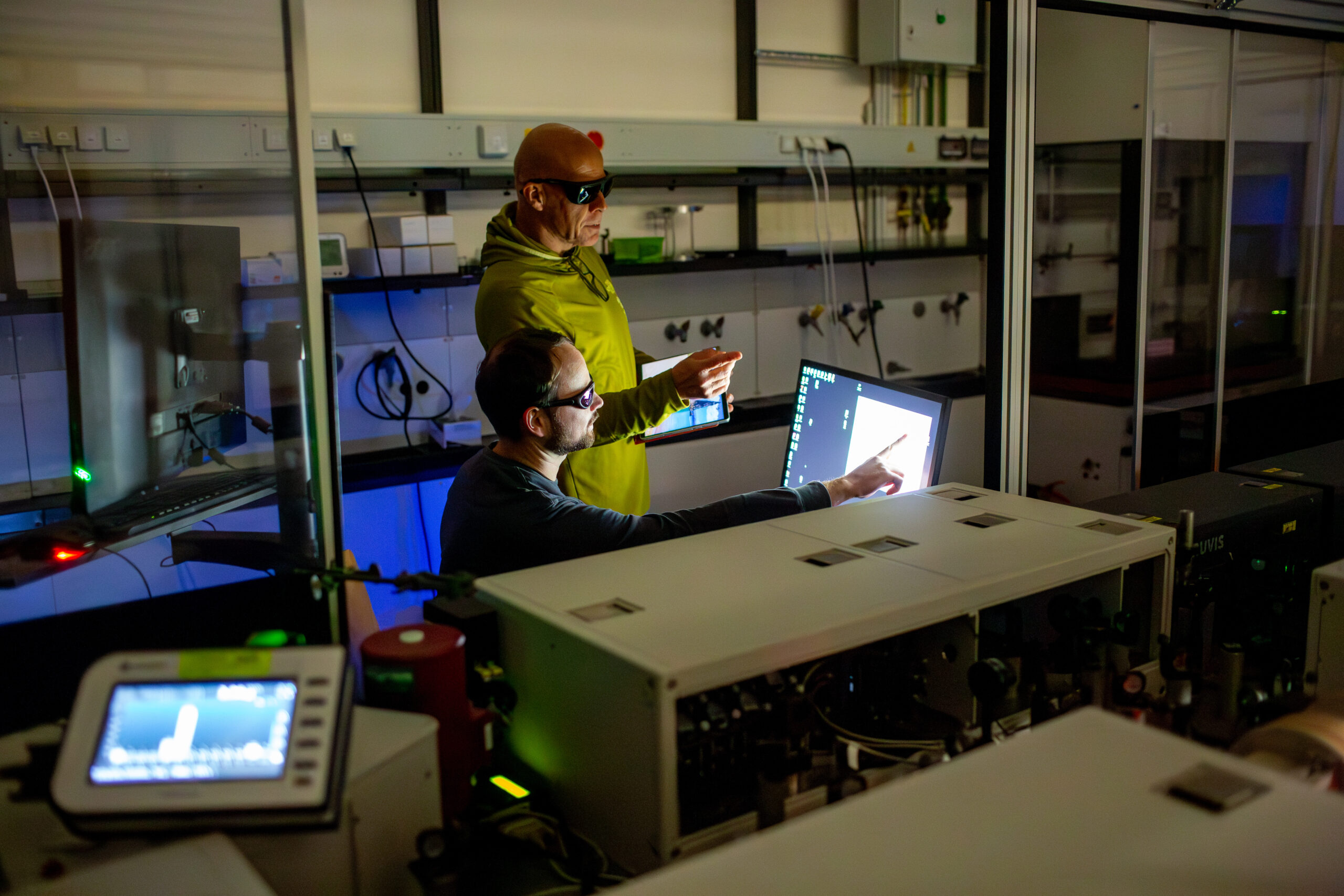 Prof. Dirk Guldi (standing) and Daniel Langford in the laser lab. Their faces are illuminated by shimmers of light. They are wearing safety goggles.