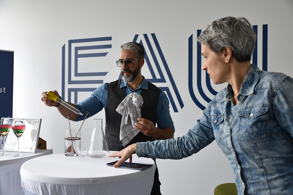 The team consists of a woman with short, mottled gray hair and a barista with short gray hair and thick-rimmed fashionable glasses. They are preparing everything for cocktail mixing.
