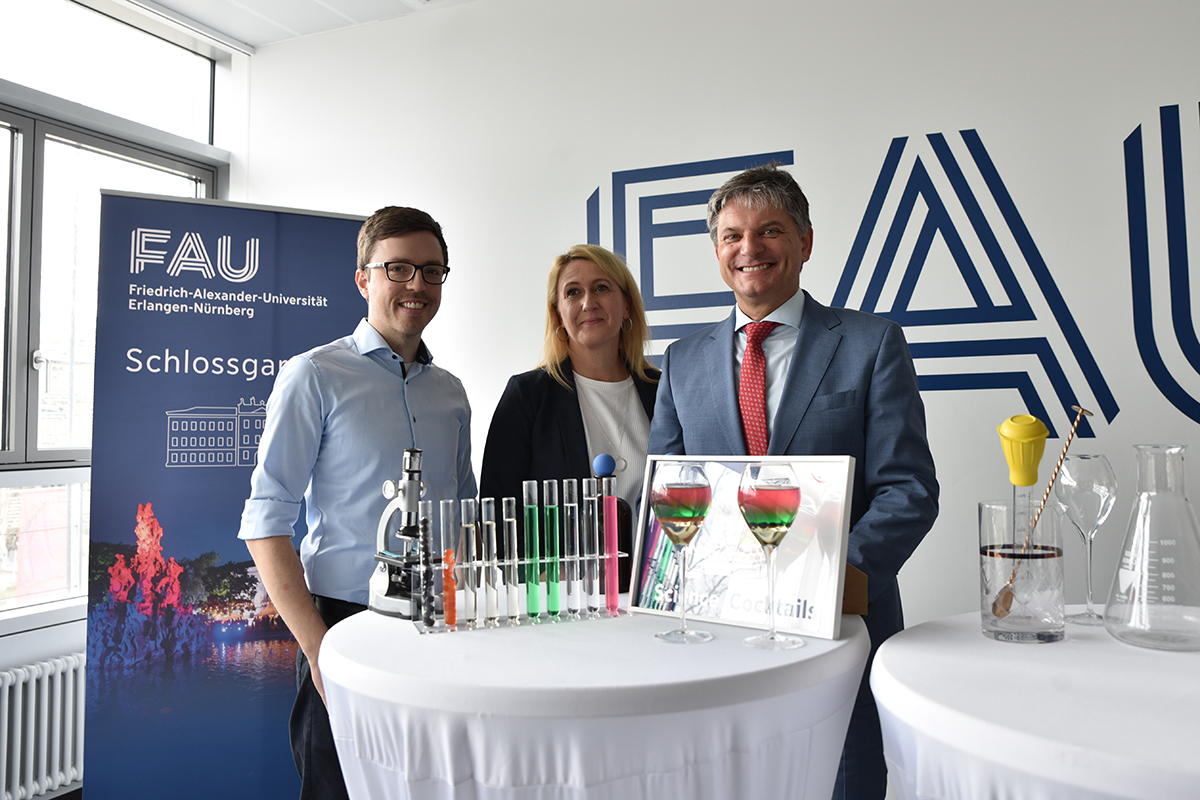 Max Gmelch, a young man with brown hair and glasses, Birgit Baumgart, a middle-aged woman with blonde hair, and President Hornegger, a middle-aged man, are standing at a high table with two cocktail glasses in front of them and test tubes filled with colored liquids next to a microscope.