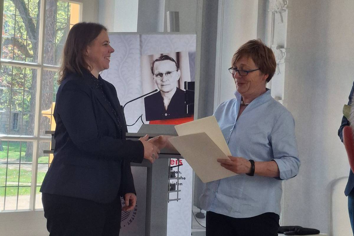 Senior Councillor Bergmann, a middle-aged woman with short hair and glasses, hands over the prize to Häneke, a younger woman with shoulder-length hair.