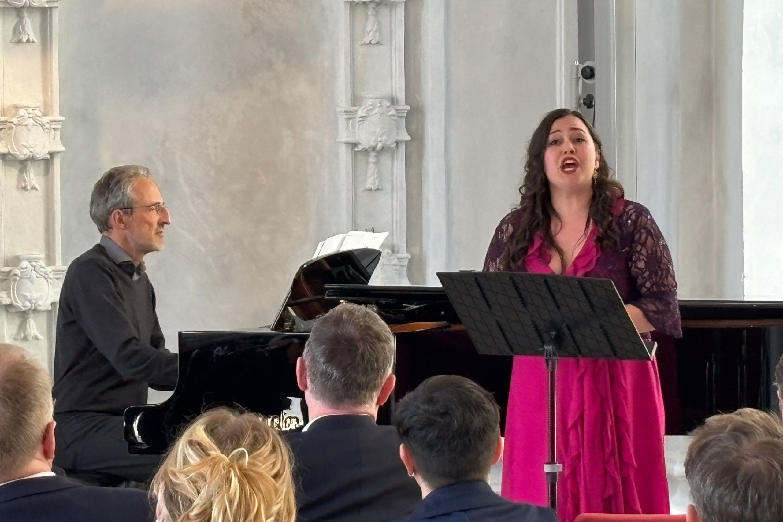 Klek, an older man with mottled gray hair on a grand piano, Eismont, a sturdy woman in a pink dress, sings.