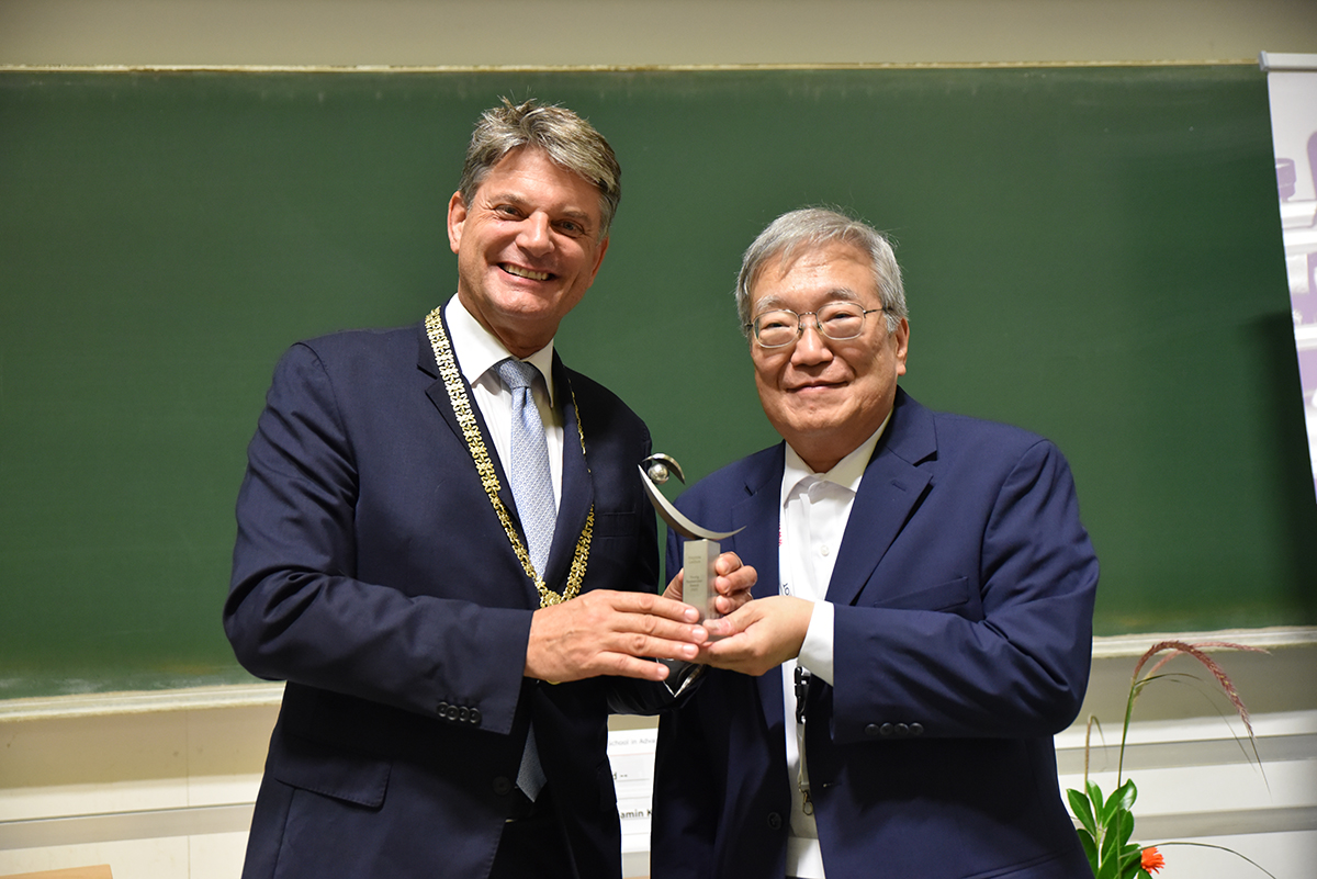 President hornegger in a dark blue suit with guest speaker Fujimoto, a gray-haired Asian, with an award of thanks