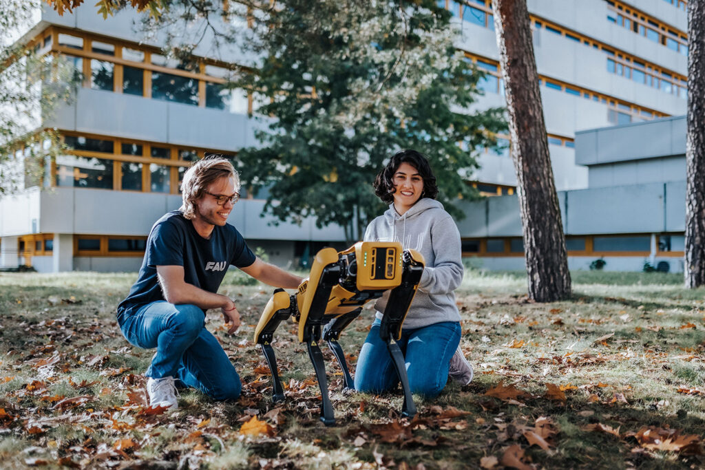 Two people are sitting with a four-legged robot on some grass on the FAU campus.