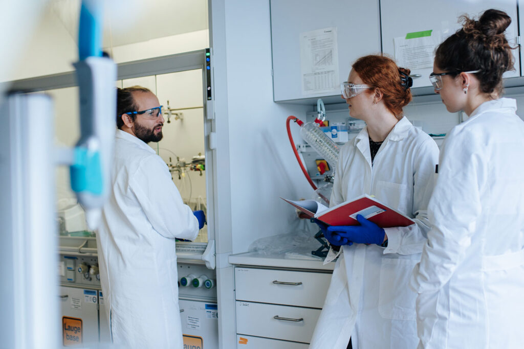 Three FAU researchers wearing lab coats, gloves and safety goggles are standing in a laboratory.