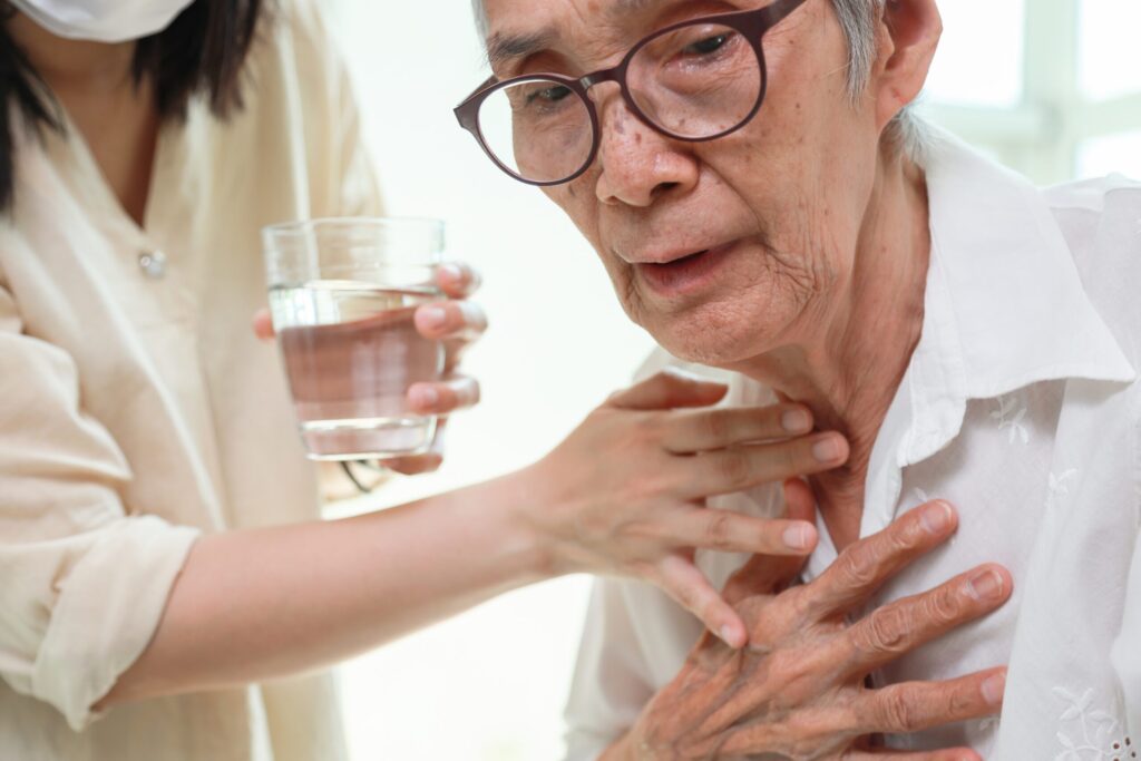 Alte Frau mit Schluckbeschwerden, im hintergrund eine Kankenschwester, die ihr helfen will und ein Glas Wasser reicht.