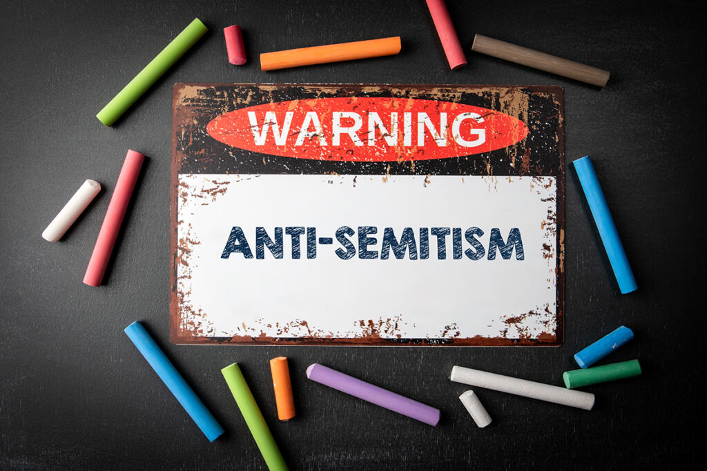 Antisemitismus. Metallisches Warnschild und farbige Kreidestücke auf einem dunklen Kreidetafelhintergrund.