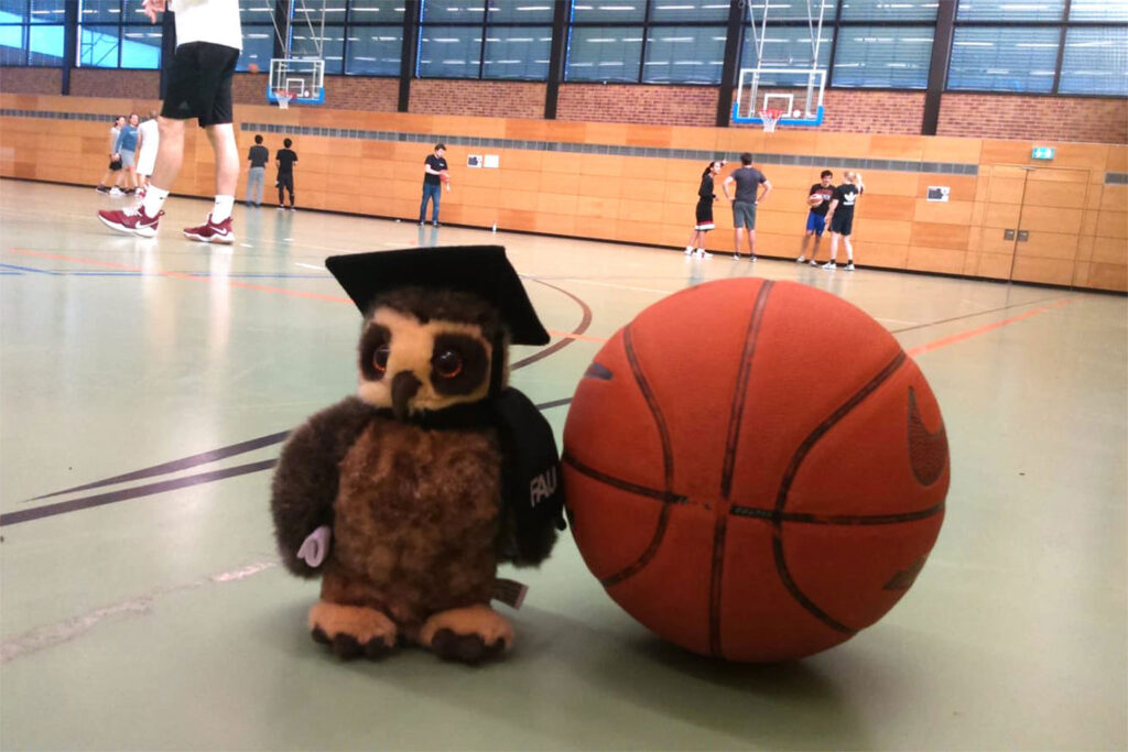 A fluffy owl and a basket ball in a sports hall. People are in the background.
