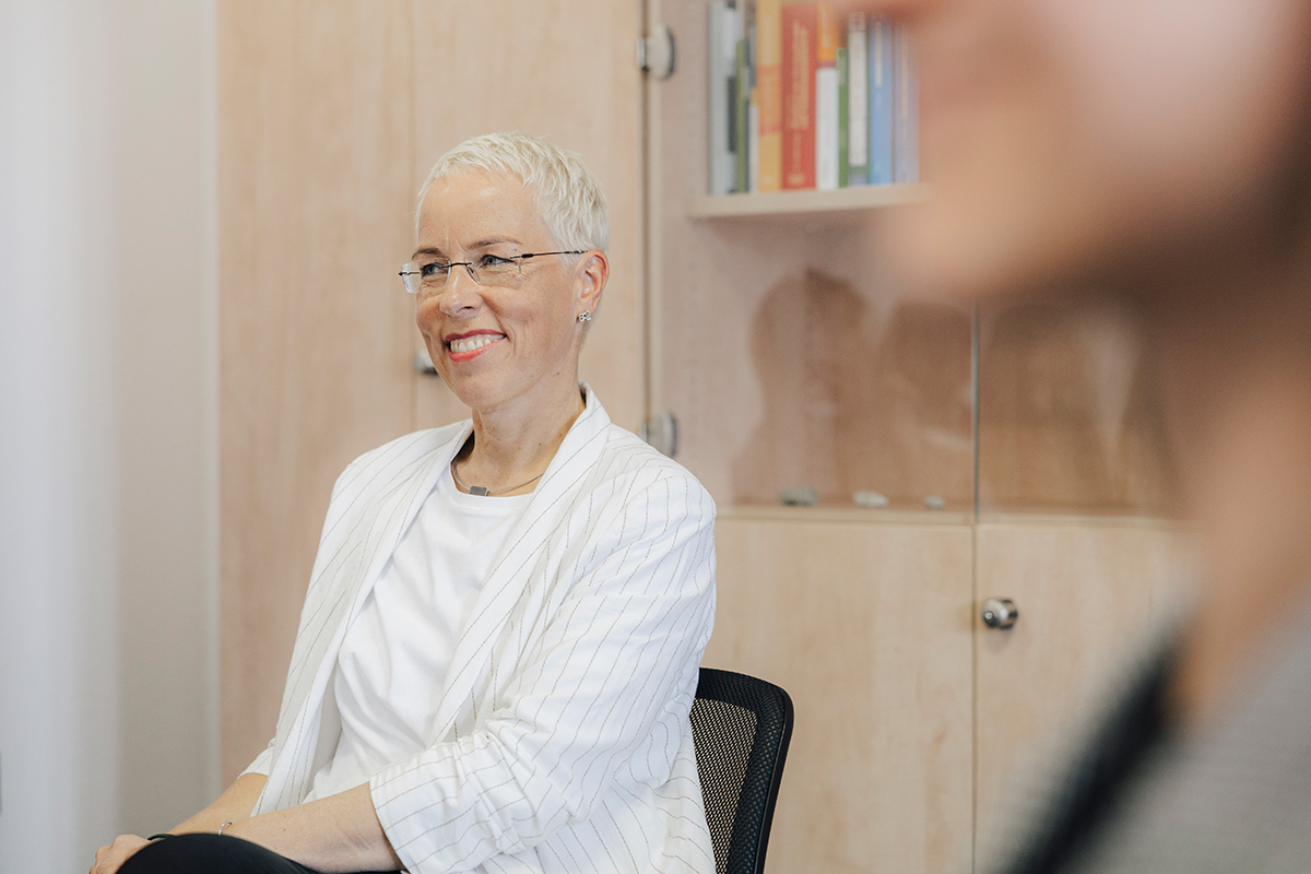 Prof. Weise sitting on a chair, smiling. The blurred outline of a person can be seen in the foreground.