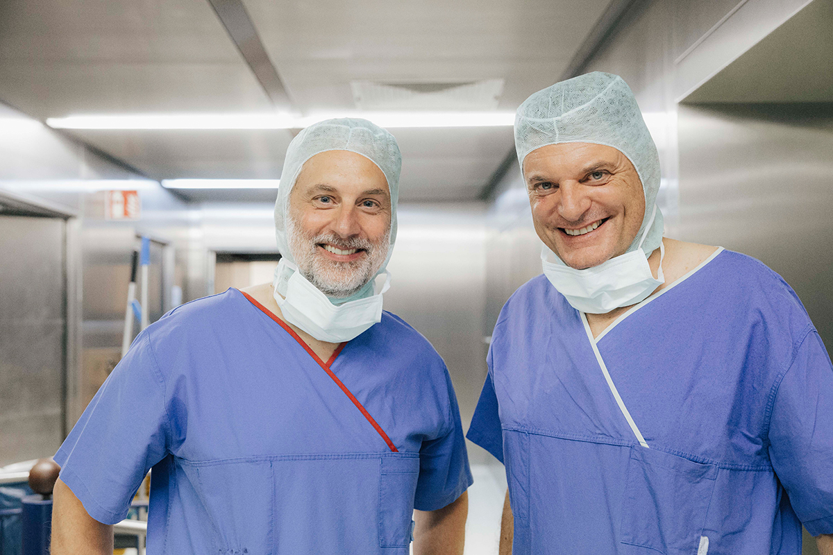 University President Hornegger and Prof. Schnell stand side by side in a hospital corridor. Both are wearing hospital scrubs, hair covers, and masks pulled down over their faces, smiling at the camera.