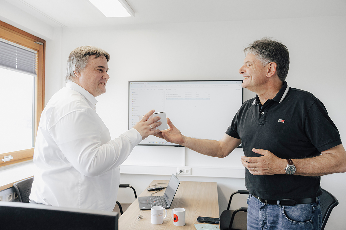 Peter Uhrig and Joachim Hornegger are standing opposite each other, Hornegger hands a small package to Uhrig.