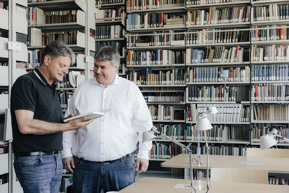 FAU President Hornegger and Prof. Uhrig are standing next to each other in a library. The President is holding a book and they are chatting.