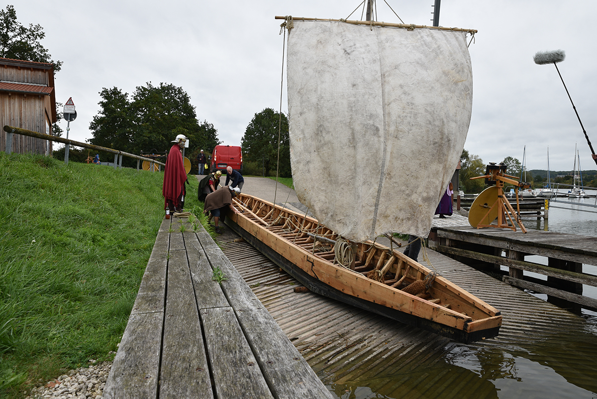 Dreyer und Helfer lassen das Boot zu Wasser, am Rande stehen Menschen in römischer Verkleidung und schauen zu.
