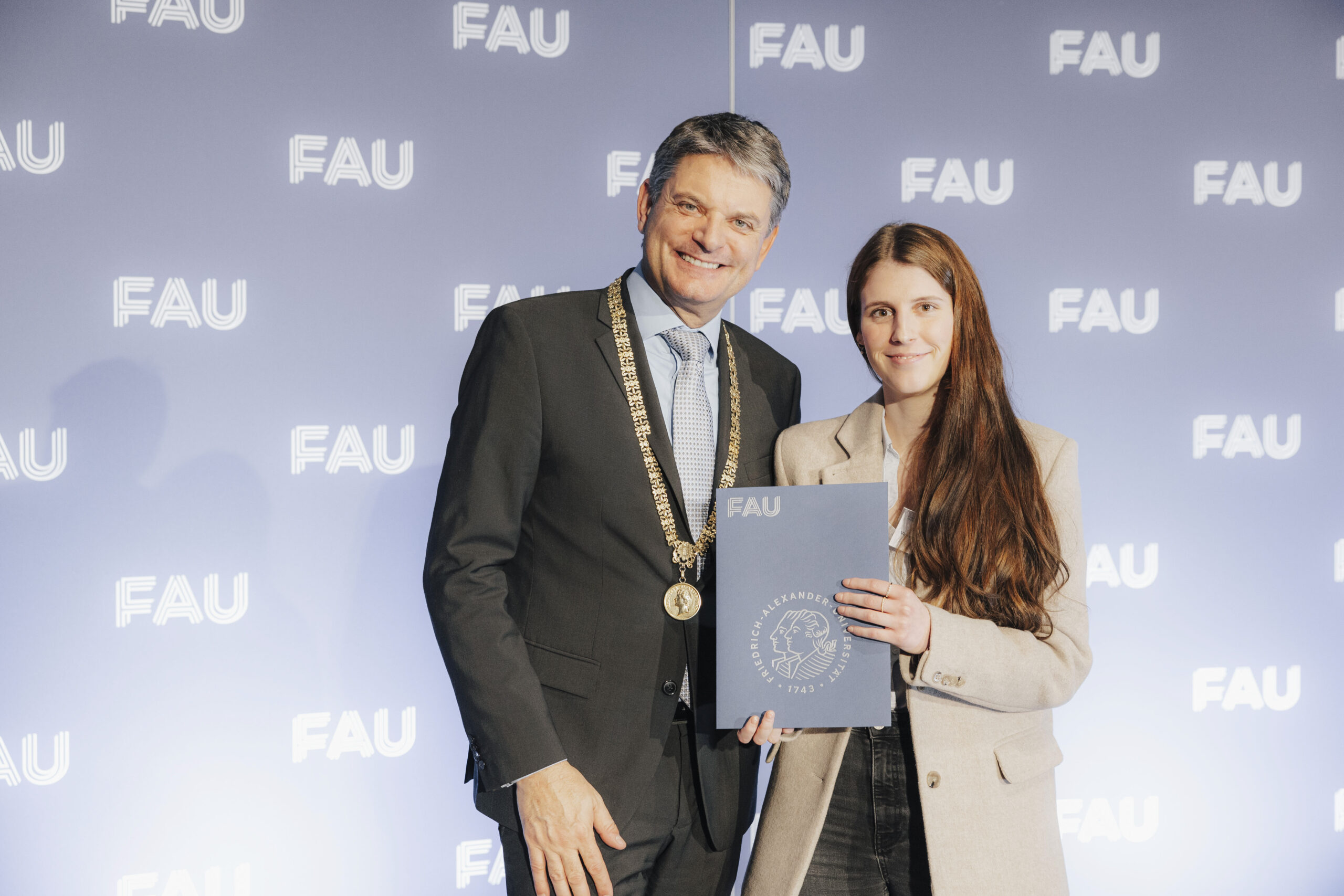 Präsident Hornegger, ein Mann in mittleren Jahren, im Anzug mit Amtskette, und Vanessa Krauss, eine junge Frau mit langen braunen Haaren, die eine Urkunde in die Kamera hält.