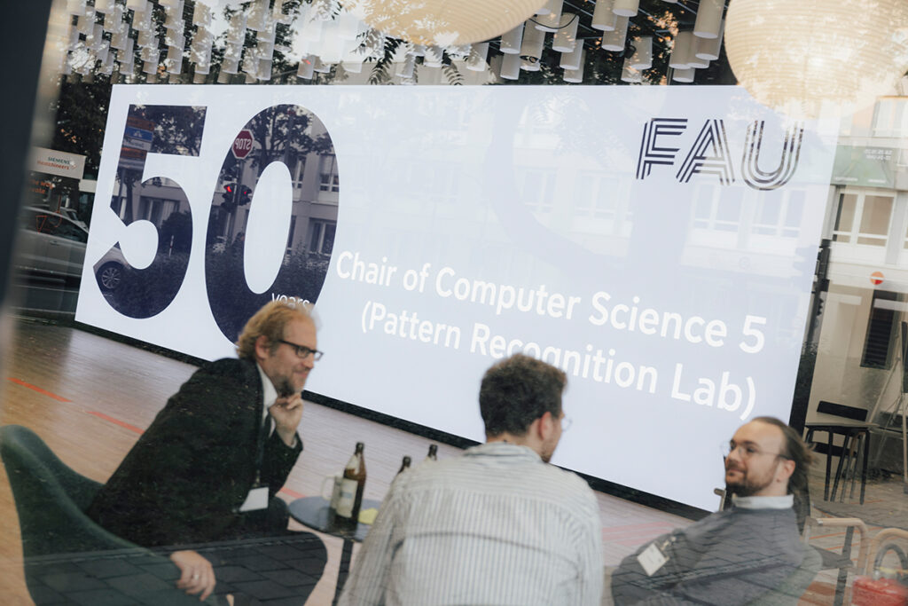 Eine Projektion mit 50 Years of Chair for Pattern Recognition. Im Vordergrund drei Männer die sich unterhalten.