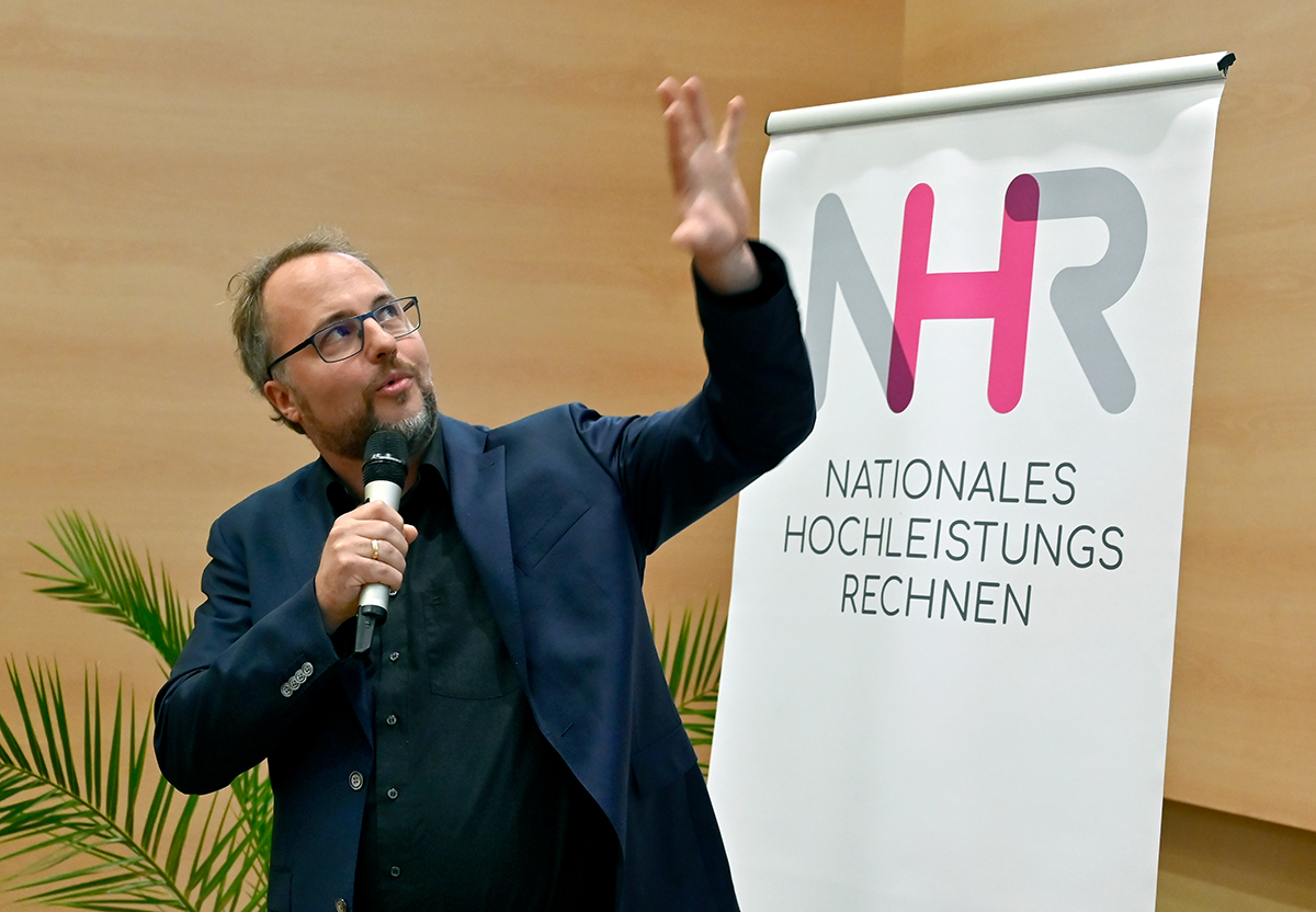 A scientist with glasses gives a speech, making a sweeping gesture and looking upwards.