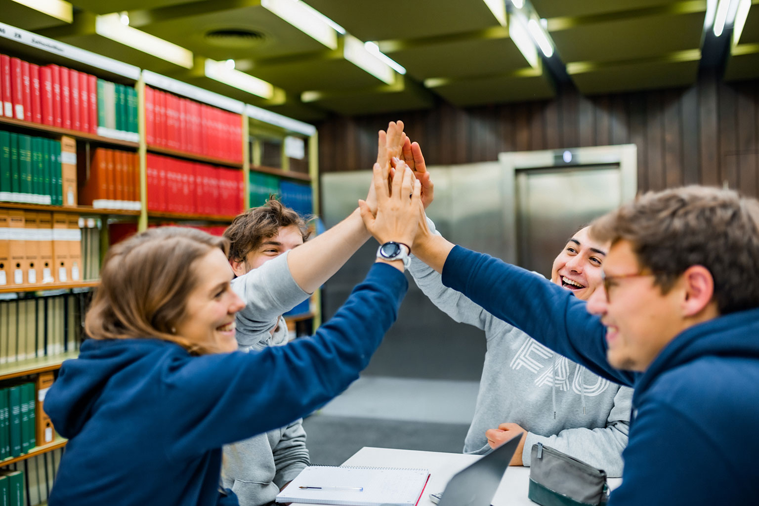 Menschen sitzten in einer Bibliothek zusammen und geben sich High-Five.