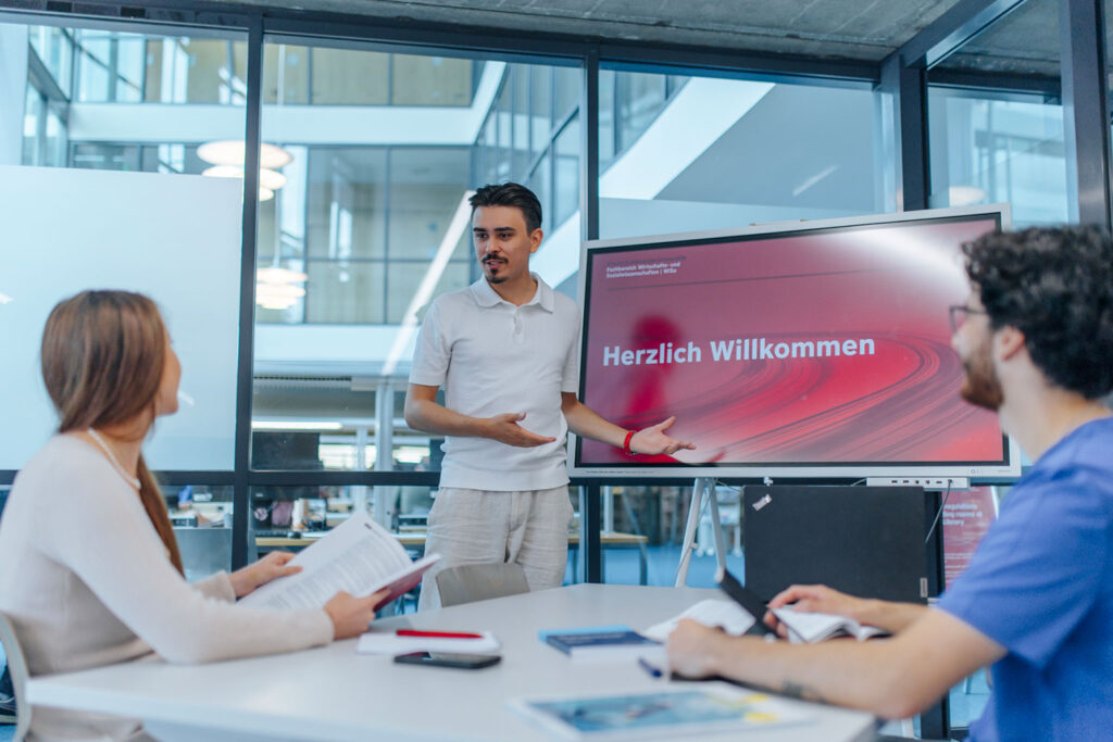 Drei Personen sitzen gemeinsam an einem Tisch vor einem großen Bildschirm. Eine der Personen hält einen Vortrag.