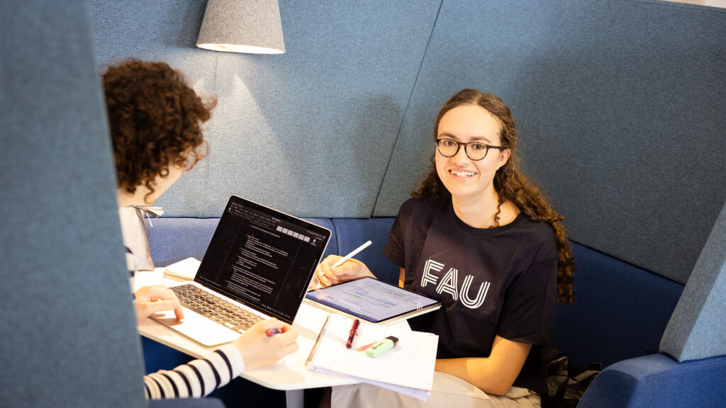 Zwei Studentinnen sitzen sich arbeitend gegenüber. Eine schaut in die Kamera und lächelt.