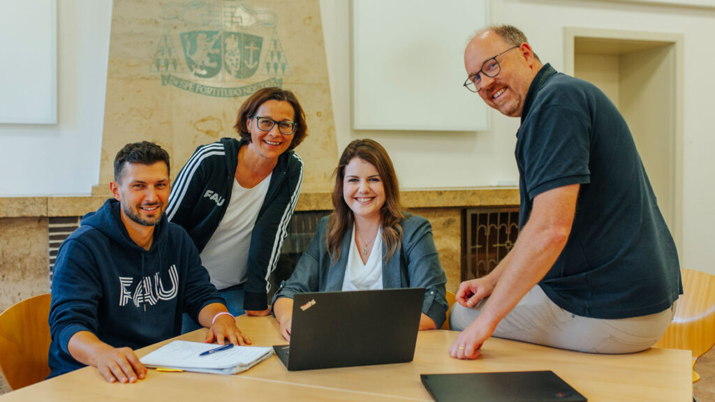 Vier Personen sitzen oder stehen um einen Laptop herum und schauen nett lächelnd in die Kamera.