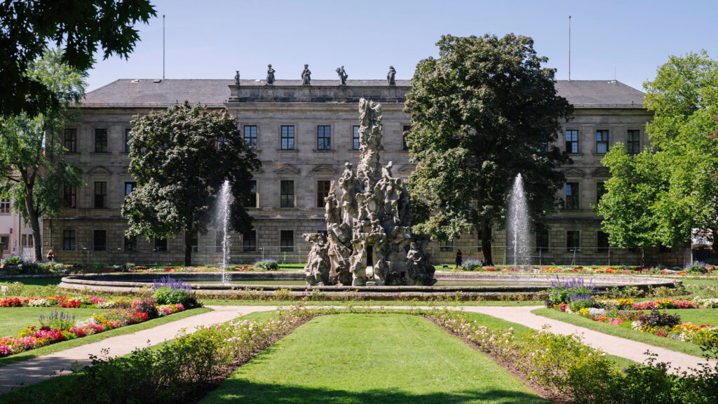 Das Schlossgebäude in Erlangen vom Park aus fotografiert. Im Vordergrund ist ein aktiver Brunnen zu sehen.
