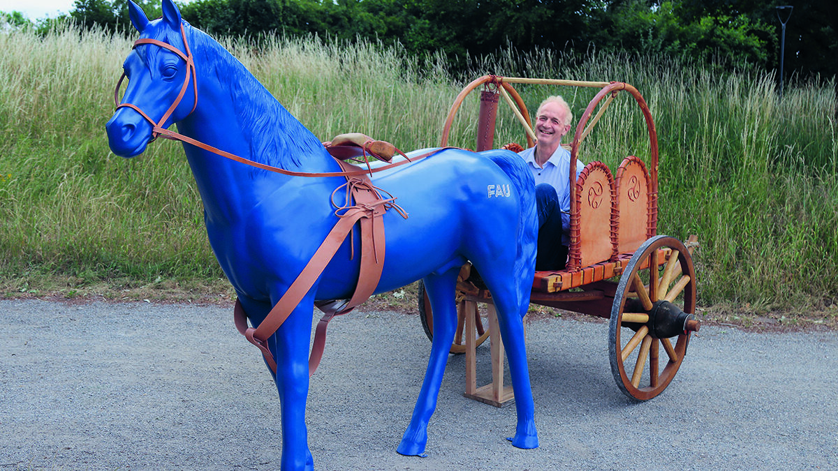 Boris Dreyer, ein schlanker Mann ende %0 sitzt lachend auf einem hölzernen Wagen vor den ein blaues Plastikpferd gespannt wurde.