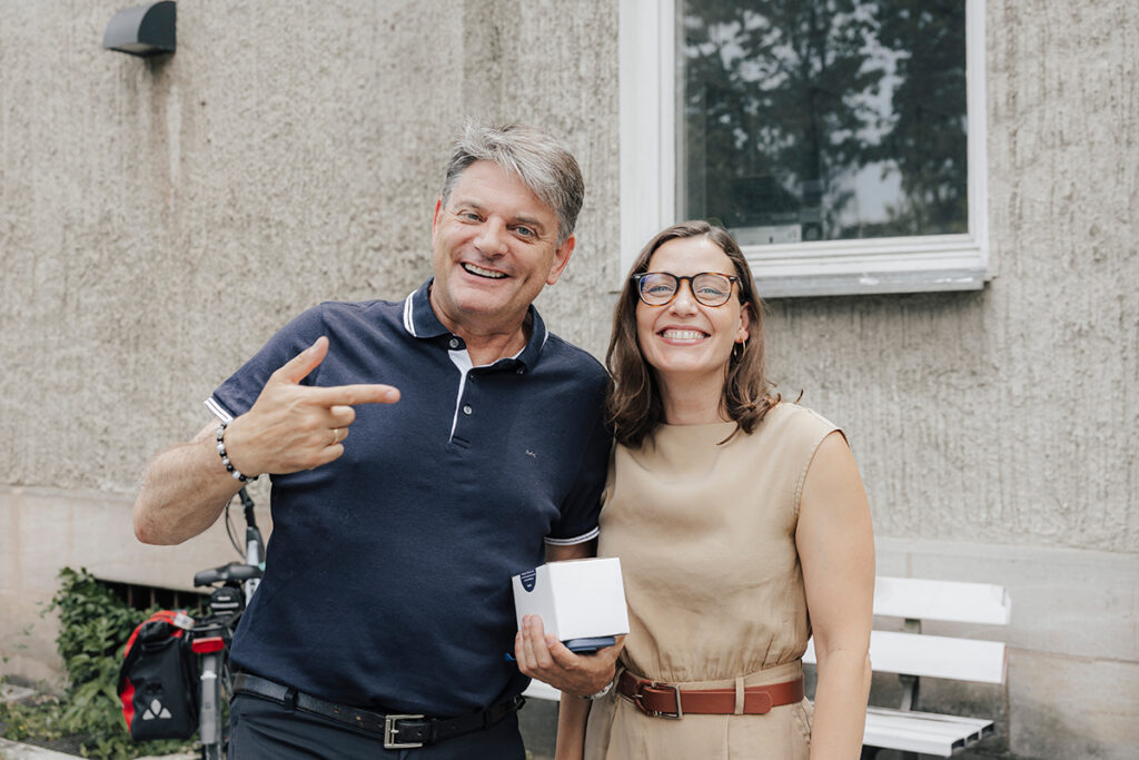 President Hornegger and Prof. Kaifie-Pechmann stand together in front of a beige house and smile at the camera. Hornegger holds a small package in his right hand and points to Kaifie-Pechmann with his left hand.