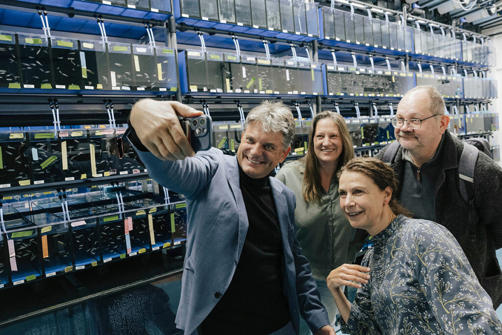 The university president, Professor Ober, and two of her colleagues pose for a selfie. Professor Hornegger holds up his smartphone, everyone looks at the camera and smiles. In the background, several wall-high rows of fish embryos are displayed.