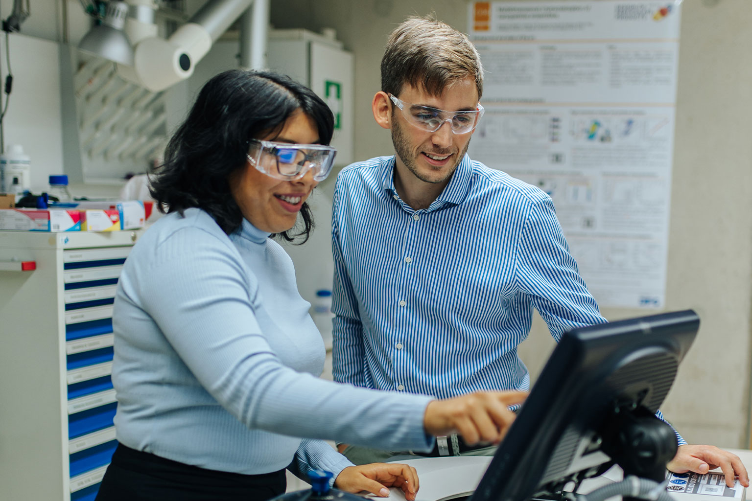 Eine Fonschende bespricht mit Ihrem Forschungsassistenten etwas an einem Monitor. Beide tragen Schutzbrillen