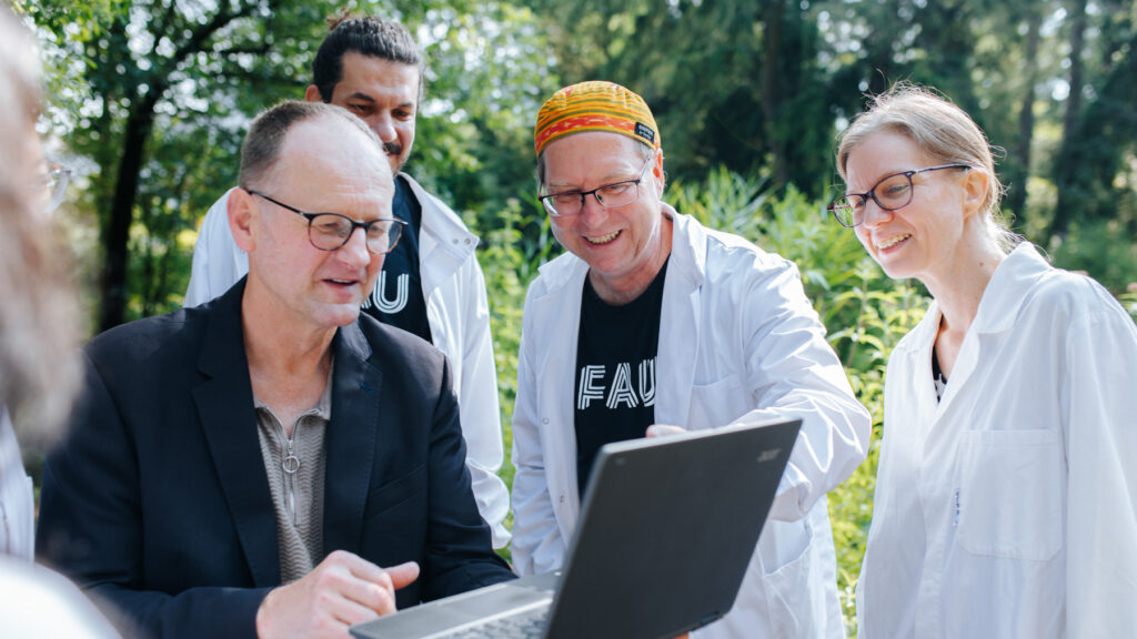 A group of researchers—some wearing white lab coats—are standing outdoors, looking at a laptop together.