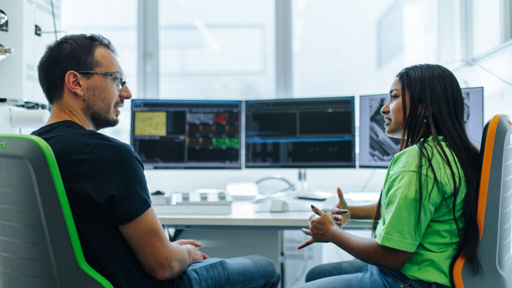 Two scientists sit in front of three screens, talking to each other.
