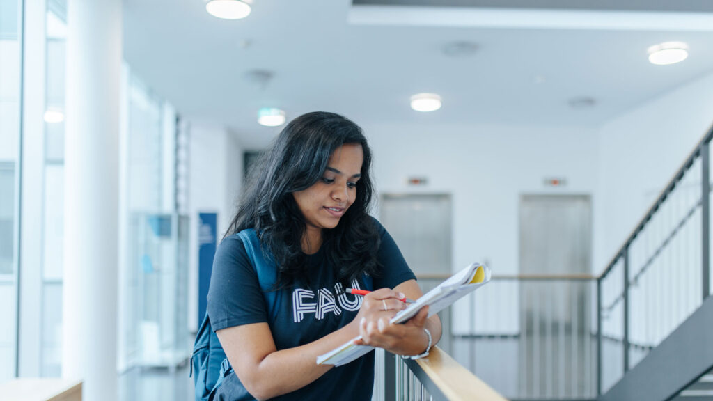 A student is leaning against a railing and writing something in a notebook.