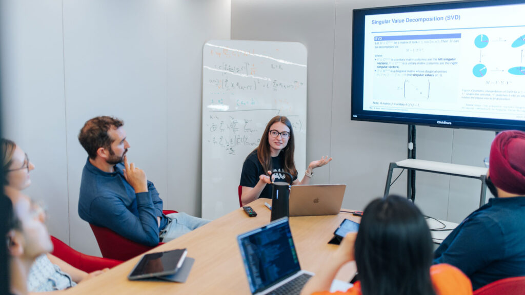 A student is giving a presentation. She is sitting in front of a screen with the instructor and other students.