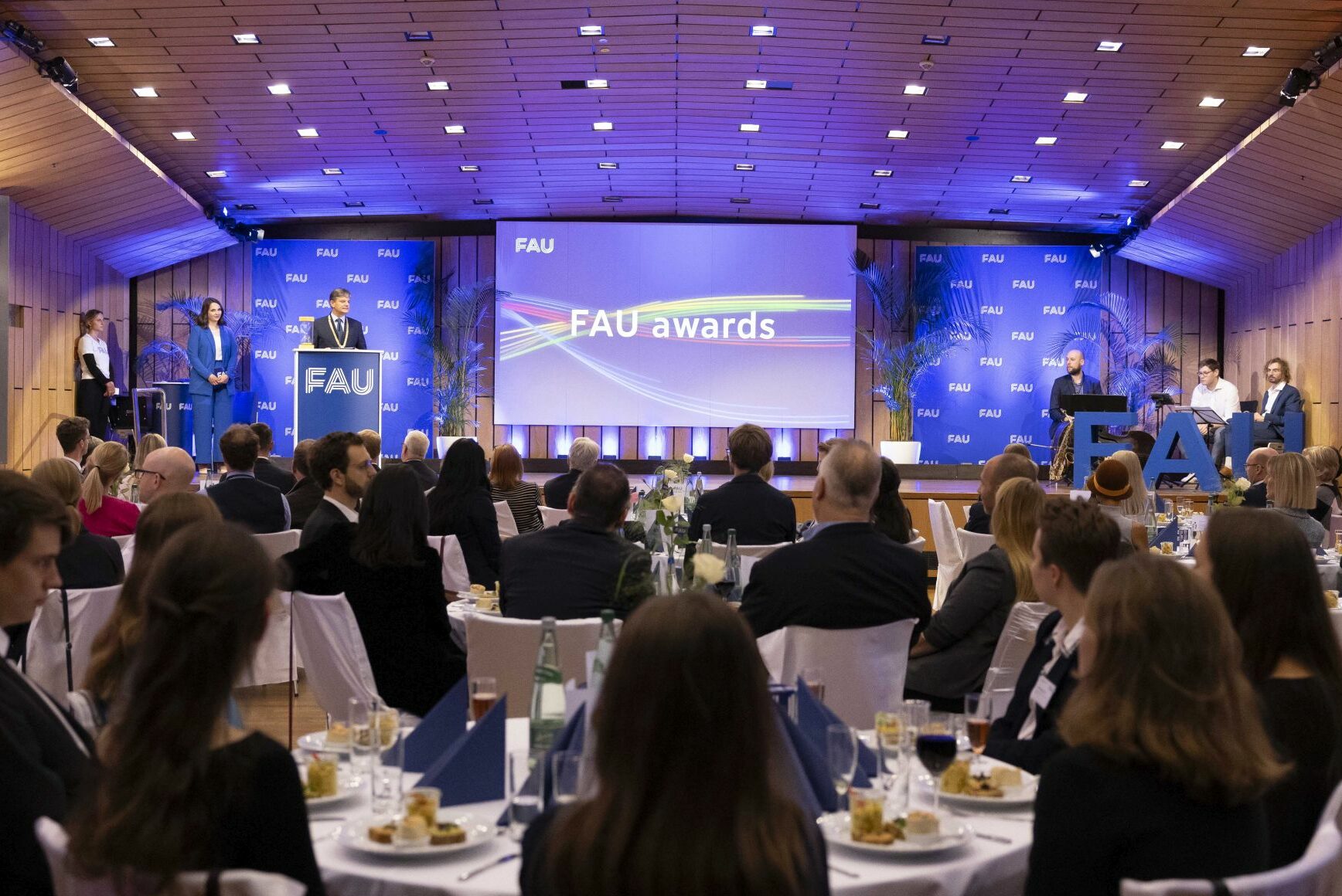 People are sitting in groups at tables, looking up at a stage. There, FAU President Joachim Hornegger is standing at a lectern, giving a speech