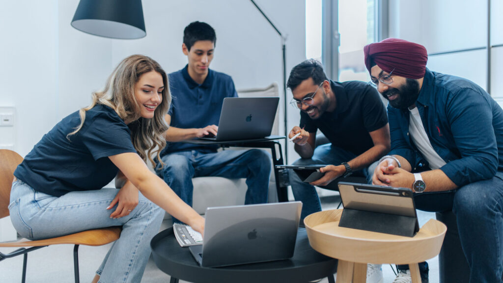 Several students are sitting together in a study café with their laptops, showing each other interesting content.