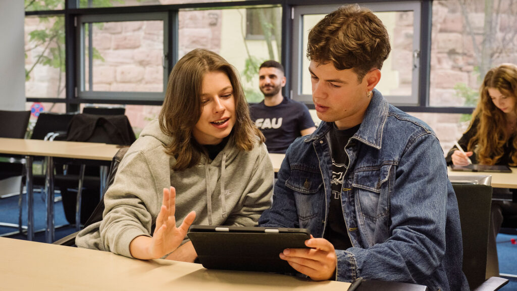 Two students sitting together in a seminar room. Both are looking into a tablet.