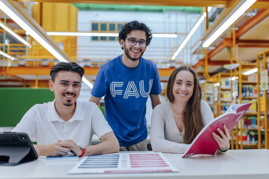 2 Studenten und eine Studentin in der in der Bibliothek, grinsen in die Kamera.