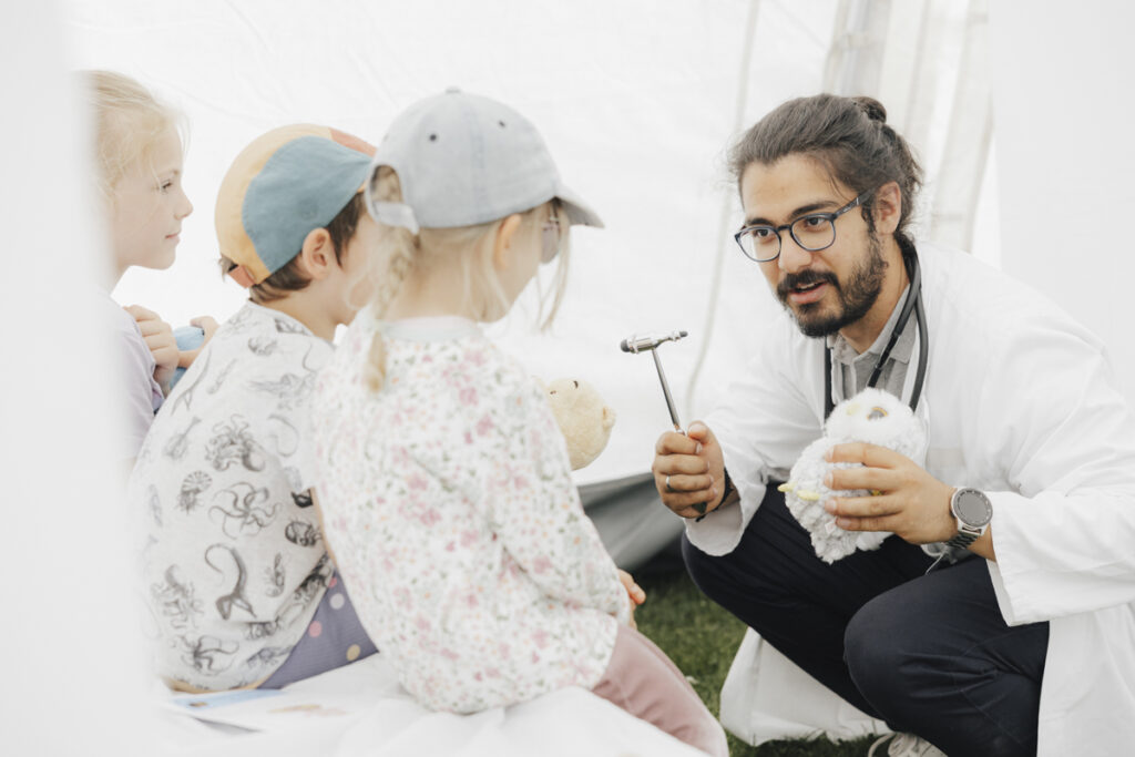 A medical student with long, dark hair and a beard explains to children using a reflex hammer