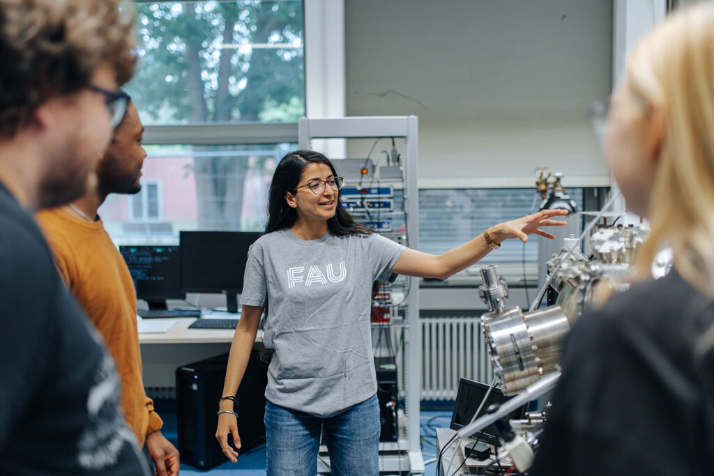 A scientists in a grey FAU-short shows other scientiests something on a machine.