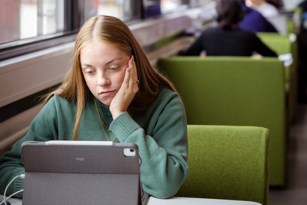 Studentin legt Gesicht in die Hand während sie in der Bib vorm Tablet sitzt