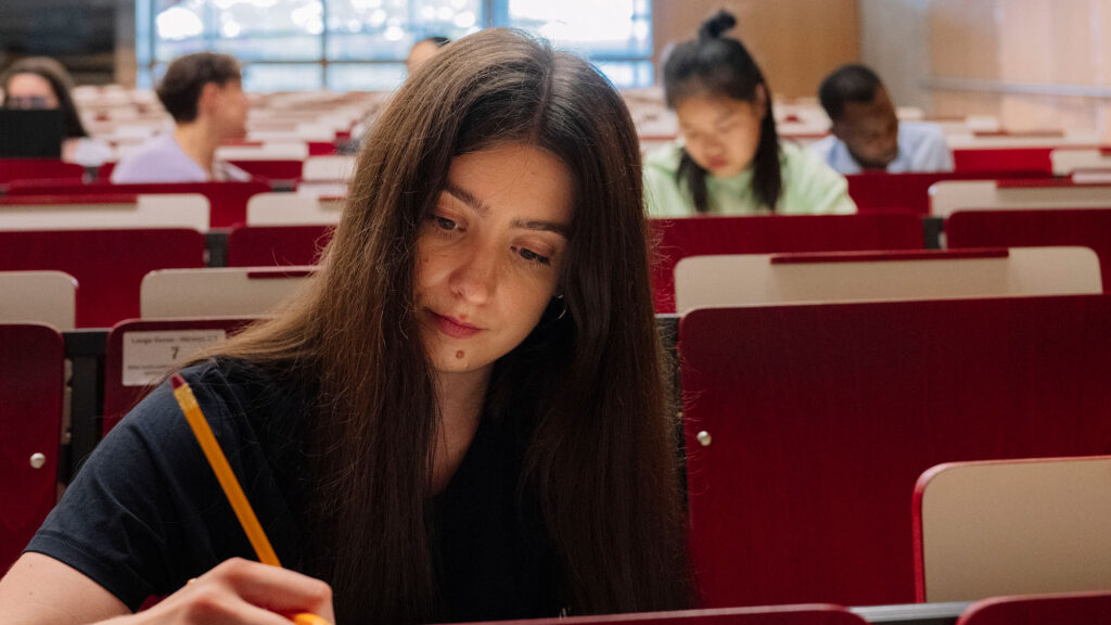 A student is sitting in a lecture hall, writing something with a pen.