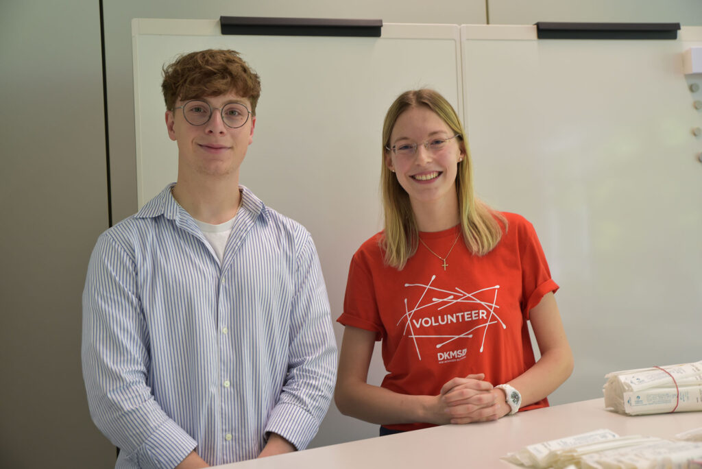 Two people smiling at the camera. The woman on the right is wearing a red DKMS top.